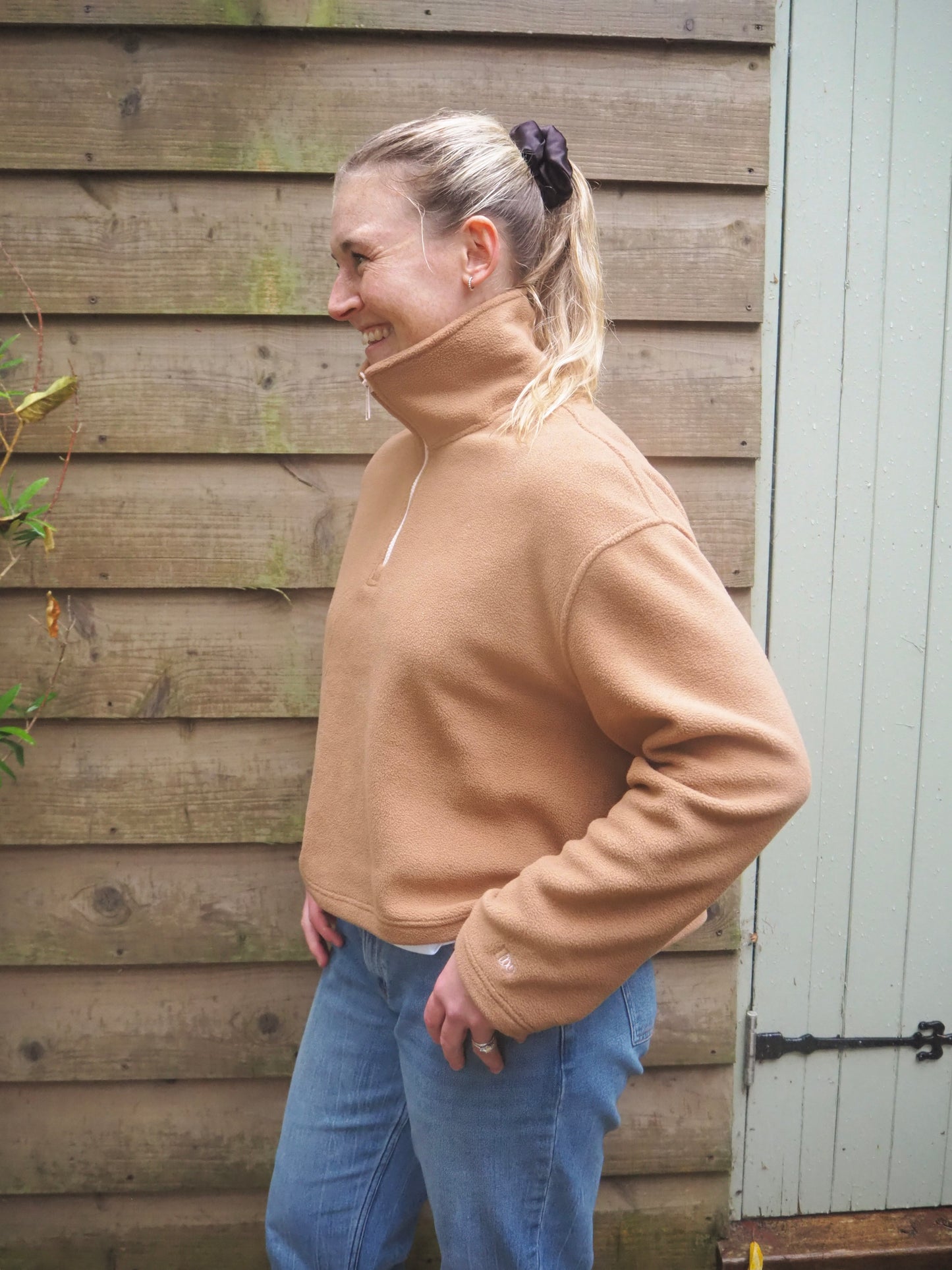 Our model Maddi wearing The Jaipur camel fleece pullover with jeans, standing outside in the countryside. 
