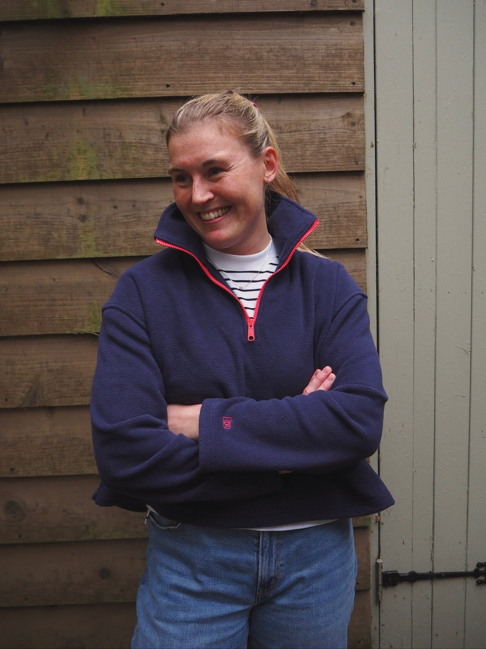Maddi is wearing the Nantucket navy blue fleece 1/4 zip pullover with a striped t-shirt underneath, standing in the countryside. 