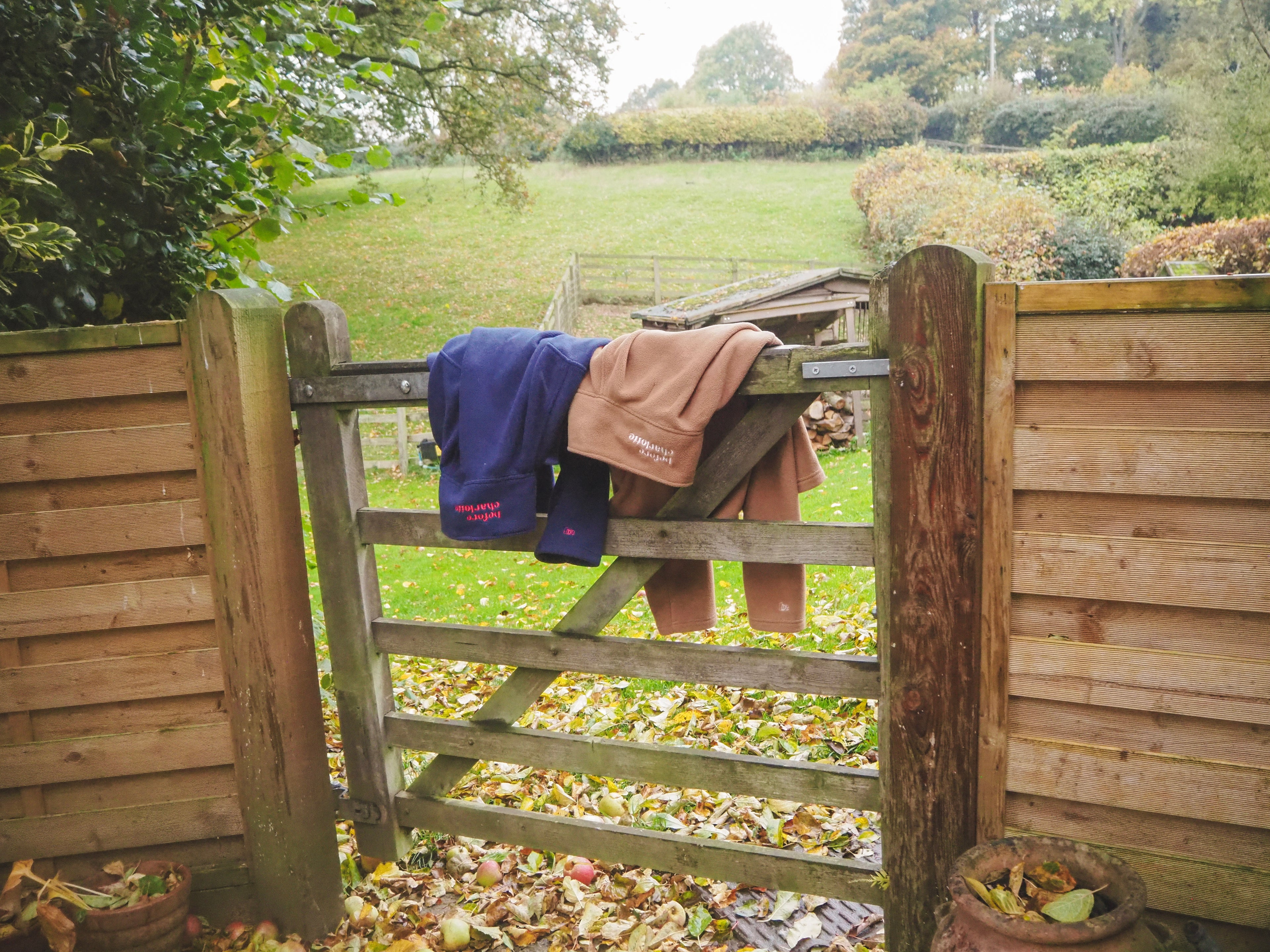 Wooden countryside gate with a blue and camel fleece draped over it, set against a grassy field. 