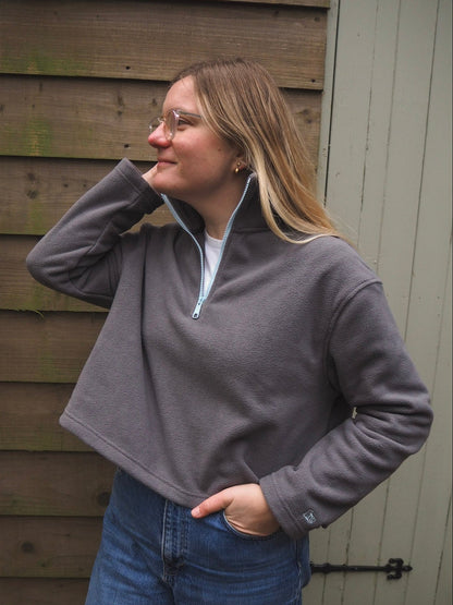 Person wearing a grey fleece pullover with a pale blue zipper, standing against a wooden wall.