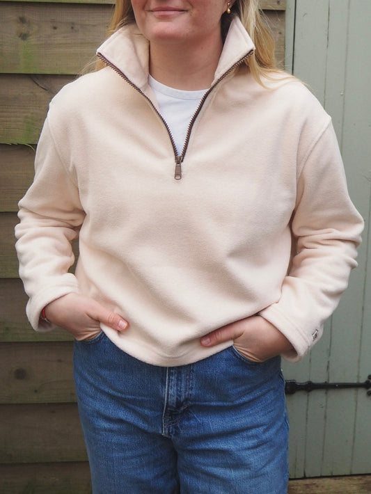 Person wearing a cream fleece pullover  fleece and blue jeans standing in front of a wooden wall.