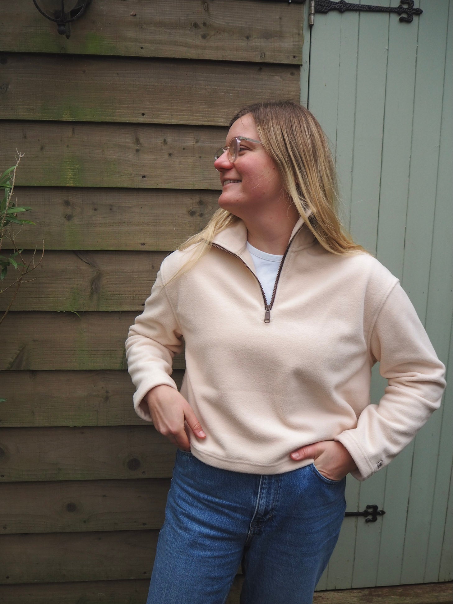 Woman wearing a cream fleece sweater and blue jeans standing against a wooden wall.