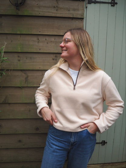 Woman wearing a cream fleece sweater and blue jeans standing against a wooden wall.